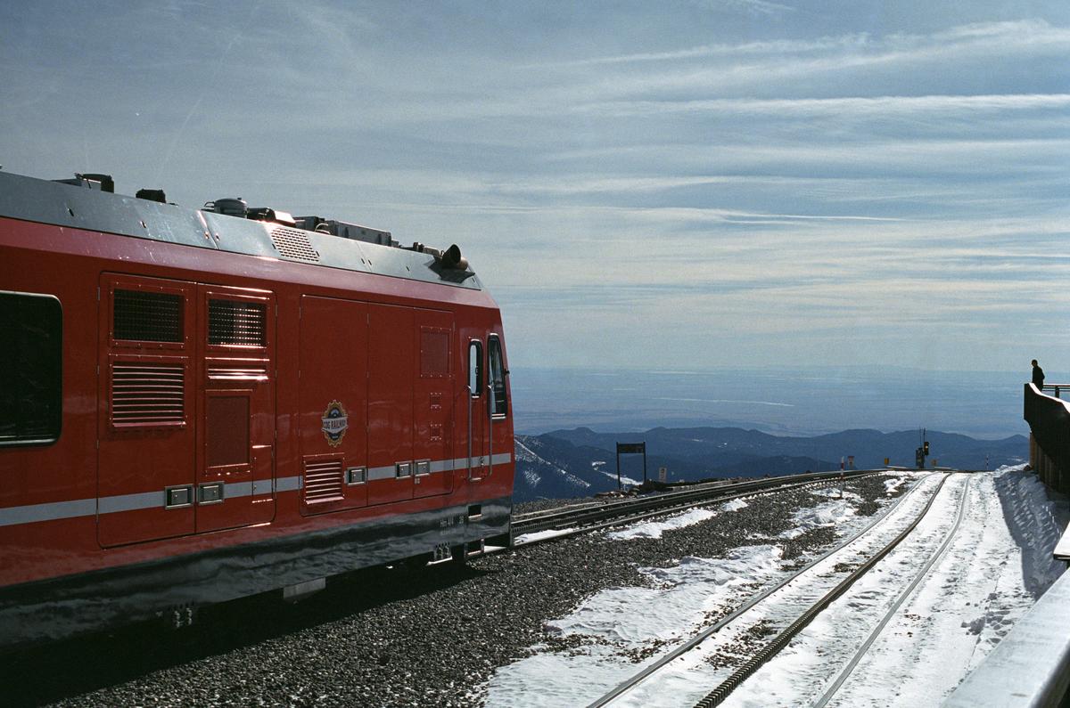 A red train engine parked in the snow at the summit of Pikes Peak, overlooking the city